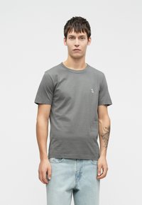 Gray cotton t-shirt with short sleeves and a crew neckline, featuring a small embroidered design on the chest. Paired with light blue jeans.