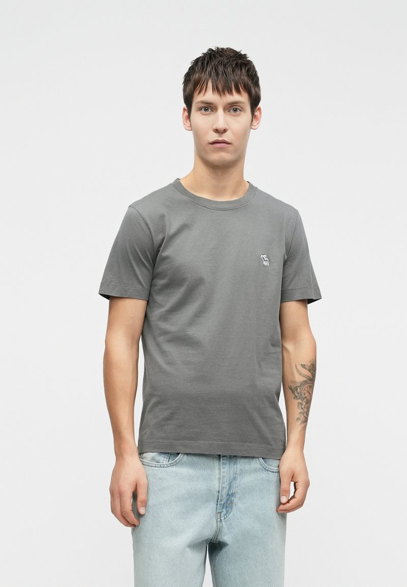 T-shirt en coton gris à manches courtes et col rond, avec un petit motif brodé sur la poitrine. Associé à un jean bleu clair.