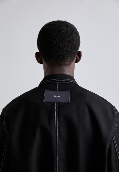 Schwarze Jacke mit kontrastierendem weißen Nähten, versehen mit einem rechteckigen Markenschild auf der Rückseite. Der Stoff wirkt robust und strukturiert.