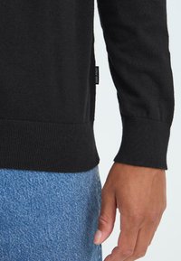Schwarzer Strickpullover mit gerippten Bündchen und einem seitlichen Etikett. Sichtbare Hand nahe dem Saum und blauer Denim-Stoff an der Taille.