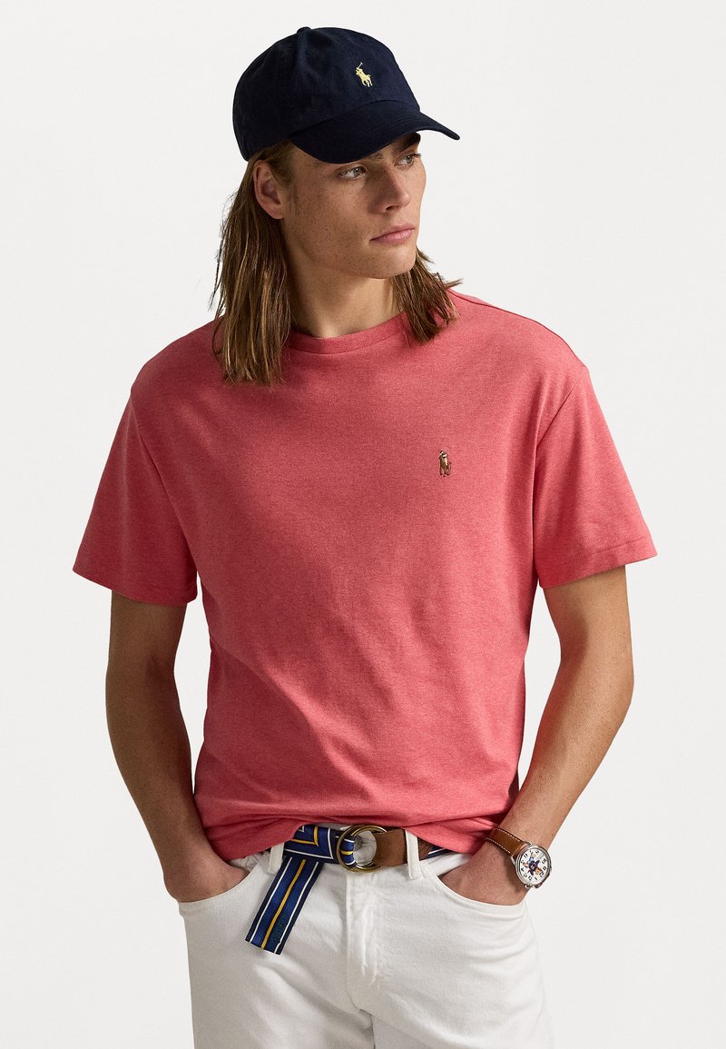 Tricou roșu din bumbac, cu mâneci scurte și un mic logo pe piept, purtat cu pantaloni din denim albi și o șapcă bleumarin.