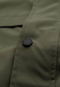 Primo piano di tessuto verde oliva con cucitura impunturata e un bottone rotondo nero etichettato con "OMBRE" più volte.