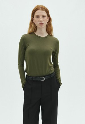 Joven mujer con cabello rojo que lleva una camiseta de manga larga verde oliva y pantalones negros con cinturón negro, de pie con las manos en los bolsillos.