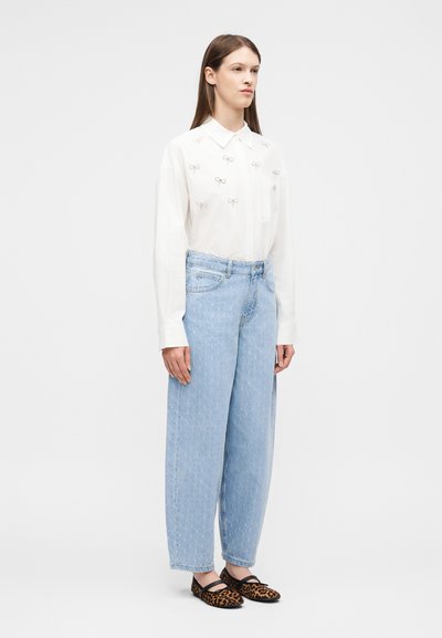 Γυναίκα που φοράει λευκό μπλουζάκι με μακριά μανίκια και μικρές λεπτομέρειες φιόγκου, ανοιχτό μπλε τζιν παντελόνι με φαρδιές γραμμές και φλατ παπούτσια με λεοπάρ σχέδιο, όρθια.