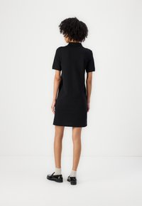 Rochie polo neagră cu mâneci scurte, croi drept, din material de bumbac, până la genunchi, cu un design simplu; asortată cu pantofi negri și șosete gri.