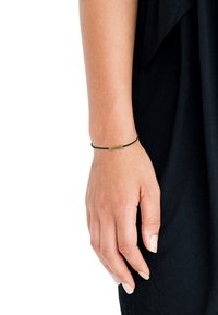 Braccialetto elastico nero con una piccola etichetta rettangolare in ottone incisa con la parola "ALWAYS". Indossato su una mano contro un indumento nero.