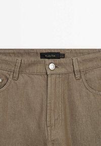 Pantalon en denim marron avec boutons à l'avant et passants de ceinture, affichant l'étiquette de la marque "Massimo Dutti" sur la ceinture intérieure.