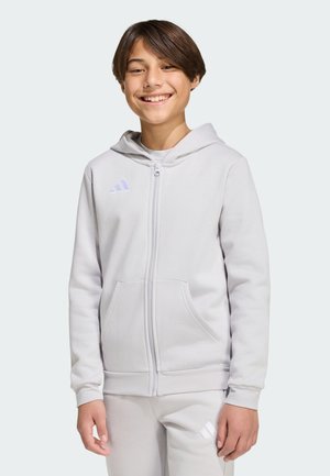 Giovane sorridente che indossa una felpa con cappuccio grigio chiaro con zip e pantaloni abbinati con loghi Adidas, in piedi davanti a uno sfondo chiaro e uniforme.