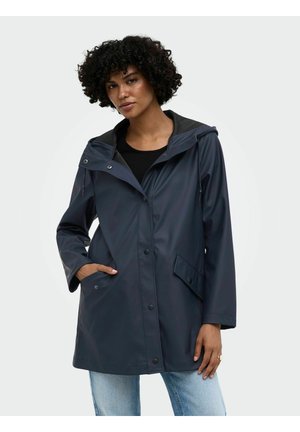 Femme aux cheveux bouclés portant un imperméable bleu marine foncé avec capuche et boutons sur le devant, associé à un jean bleu clair sur un fond uni.