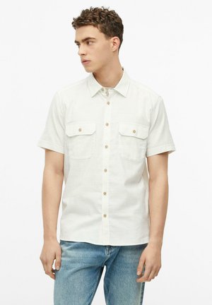 Junger Mann mit lockigem Haar, der ein weißes kurzärmeliges Hemd und hellblaue Jeans trägt und nach links schaut.