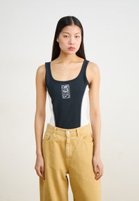 Black and white one-piece swimsuit with a front logo, featuring a sleek design and smooth texture, paired with high-waisted beige trousers.