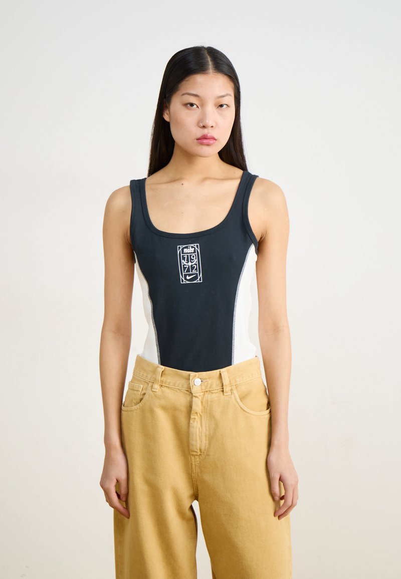 Black and white one-piece swimsuit with a front logo, featuring a sleek design and smooth texture, paired with high-waisted beige trousers.