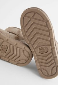 Hellbraune Wildlederstiefel mit strukturierten Gummisohlen, die ein Zickzackmuster und ein geprägtes Markenlogo aufweisen, aus der Unteransicht dargestellt.