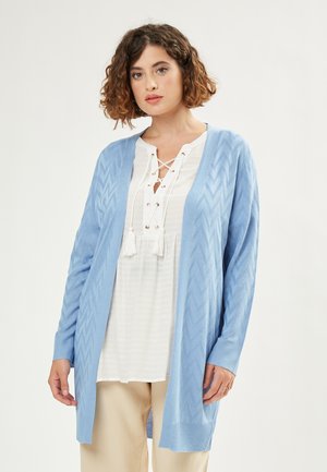 Light blue cardigan with a chevron pattern, open front, and ribbed cuffs. Paired with a white lace-up top and beige trousers.