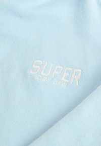 Tessuto azzurro chiaro con una texture morbida, caratterizzato da un ricamo in bianco con la scritta "SUPER DRY-JPN" in bella vista.