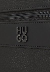 Material texturat negru, cu un fermoar și un logo „HUGO” ridicat din metal negru lucios. Designul este minimalist și elegant.