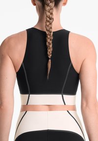 Crop top de sport noir avec une bande crème contrastante, présentant des détails de couture et un design dos nageur. La vue arrière montre une coiffure tressée.