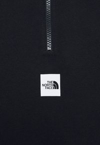 Tissu noir avec une courte fermeture éclair verticale au-dessus d'une étiquette carrée blanche affichant le logo The North Face en noir.