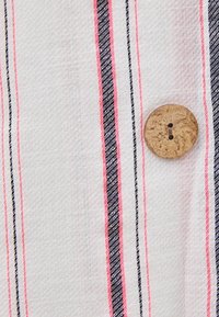 Bomullstyg med vit bakgrund, med rosa och marinblå vertikala ränder. En rund, naturlig träknapp med två hål syns.