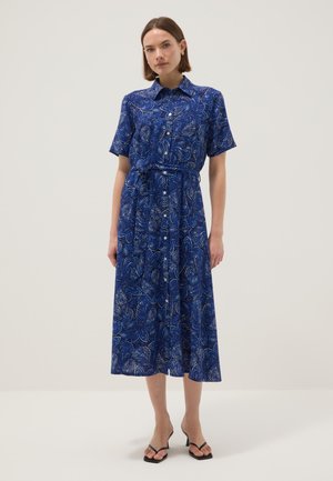 Femme portant une robe midi bleue à motifs avec boutons, manches courtes et sandales noires à talons, debout devant un fond uni.