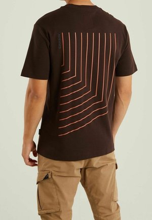 Hombre con camiseta marrón oscuro con líneas rojas verticales y en ángulo en la espalda, combinado con pantalones cortos cargo color beige, de pie contra un fondo liso.