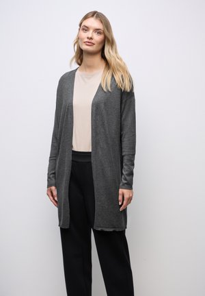 Femme aux longs cheveux blonds portant un haut beige, un cardigan ouvert gris foncé et un pantalon noir, debout devant un fond clair uni.