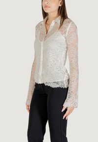 Blusa in pizzo color avorio chiaro con colletto, chiusura a bottoni e maniche lunghe. Design floreale trasparente con dettagli a scallop sull'orlo.