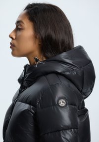 Chaqueta acolchada negra con un acabado brillante, cierre de cremallera y capucha. Incluye un parche con el logo en la manga y un cordón ajustable en el cuello.