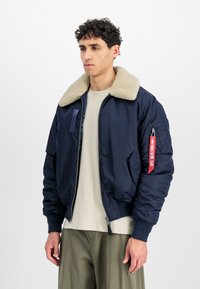 Marineblå bomberjakke med en beige shearling-krave, lynlås lukning og rød stofmærke som accent. Glat materiale og ribbede manchetter.