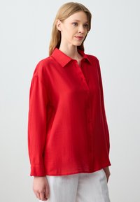 Red button-up shirt with a collar, long sleeves, and a relaxed fit. Made from a textured fabric, featuring a smooth finish and front buttons.