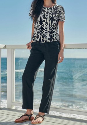 Kobieta ubrana w czarno-białą bluzkę w krótkim rękawie w wzory, czarne spodnie i czarne sandały, stoi na pokładzie przy barierce nad oceanem.