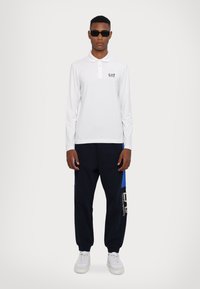 Hvit langermet polo med krage og EA7-logo, sammen med marineblå joggebukser med blå detaljer og logo-print, komplett med hvite sneakers.