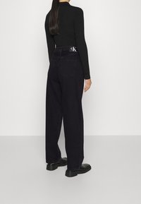 Mujer de pie con la espalda hacia la cámara, vistiendo un suéter negro de canalé con cuello de tortuga, jeans oscuros de pierna ancha con el logo de CK, y zapatos de plataforma negros.