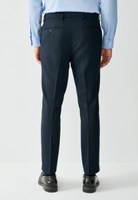 Donkere blauwe pantalon in gladde stof met een getailleerde pasvorm, voorzien van achterzakken en een plooi in elke pijp, gecombineerd met zwarte schoenen.