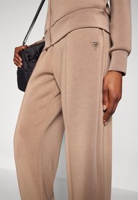 Pantalons beige à jambes larges en tissu lisse, avec un détail de logo triangulaire sur la hanche et des poches latérales pour plus de fonctionnalité.