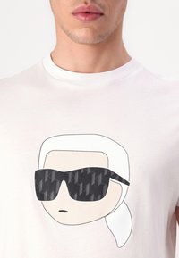 Bílé bavlněné tričko s grafikou postavy s bílými vlasy a černými slunečními brýlemi, které mají geometrický vzor.