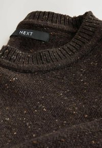 Dunkelbrauner gestrickter Pullover mit geripptem Kragen. Textil mit einer strukturierten Oberfläche und helleren Farbnuancen. Das Etikett trägt die Aufschrift "NEXT".