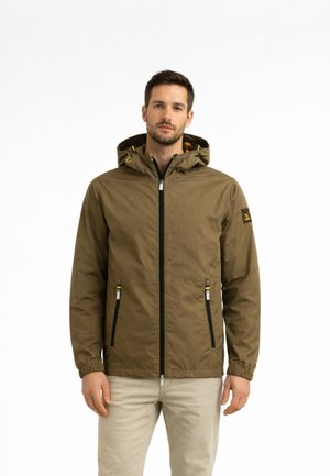 Uomo che indossa una giacca marrone con cappuccio, cerniera nera e tasche, in piedi davanti a uno sfondo bianco uniforme.