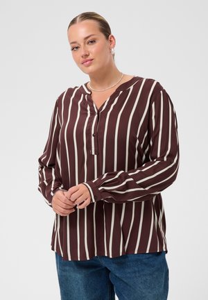 Femme portant un chemisier marron à manches longues avec des rayures verticales blanches et un jean bleu, debout les mains jointes devant elle.