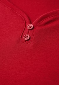 Červená bavlněná košile s výstřihem do V, dvěma červenými knoflíky na krku a hladkým, texturovaným materiálem.