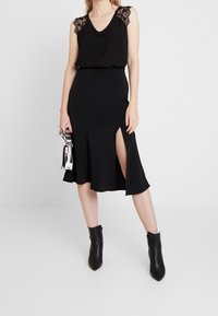 Nainen, joka on pukeutunut mustaan hihattomaan paita, jossa on pitsireunus, mustaan midi-hameeseen, jossa on sivuhalkio, mustiin nilkkureihin ja pitää käsissään hopeista käsilaukkua.