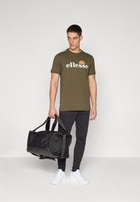 Olive green t-shirt with white "ellesse" logo, paired with black sweatpants and white sneakers. Man holds a black duffel bag.