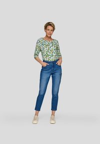 Langärmliges Oberteil mit Blumenmuster in Grün und Blau, kombiniert mit engen blauen Jeans mit Verzierungen. Das Model steht mit den Händen in den Taschen.