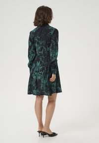 Schwarzes Kleid mit grünem Blumenmuster, langen Ärmeln und einem ausladenden Rock. Glattes Material mit leicht glänzender Oberfläche. Offene Rückenpartie.