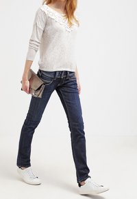 Top de manga larga gris claro con escote de encaje, combinado con jeans ajustados azul oscuro y zapatillas blancas. Sostiene un clutch metálico.