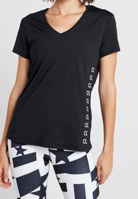 Kobieta w czarnej koszulce z krótkim rękawem i dekoltem w serek z logo po boku oraz legginsach w czarno-biały geometryczny wzór, stojąca na jednolitym tle.