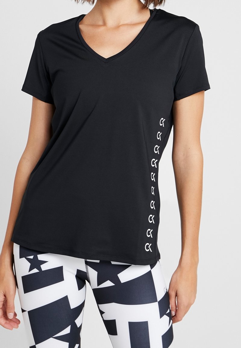 Kobieta w czarnej koszulce z krótkim rękawem i dekoltem w serek z logo po boku oraz legginsach w czarno-biały geometryczny wzór, stojąca na jednolitym tle.