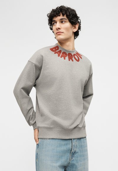 Νέος άνδρας με σγουρά μαλλιά, φορώντας γκρι φούτερ με κόκκινο κείμενο "BARROW" κοντά στον γιακά και ανοιχτόχρωμα μπλε τζιν, κοιτάζει προς τα πλάγια.