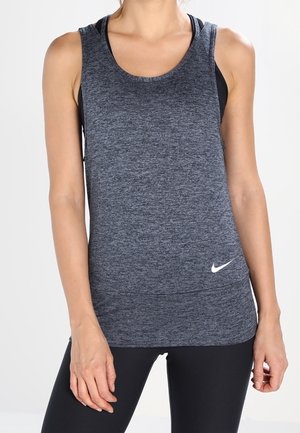 Femme portant un débardeur de sport gris foncé sans manches avec un logo Nike blanc et des leggings noirs, debout devant un fond uni.
