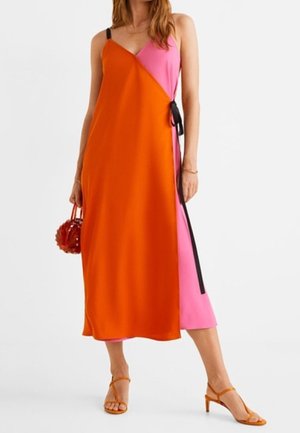 Kvinde iført en ærmeløs midi-kjole i farveblokke af orange og pink med sort sidesløjfe, orange hælede sandaler og med en rød rund håndtaske.